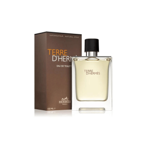 A high-angle shot of a bottle of Hermès Terre d'Hermès Eau de Toilette next to its brown rectangular box. The glass bottle is transparent with a thick base, and the fragrance liquid inside is a light amber color. The brand name and product title are printed in white on the bottle and in a slightly darker amber on the box. The cap is black. The background is white.