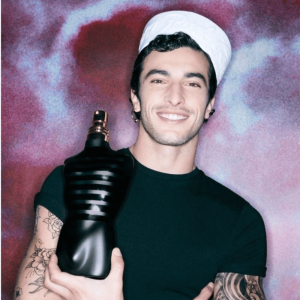 A smiling man with tattoos and a sailor hat holding a bottle of Jean Paul Gaultier Le Male Le Parfum.